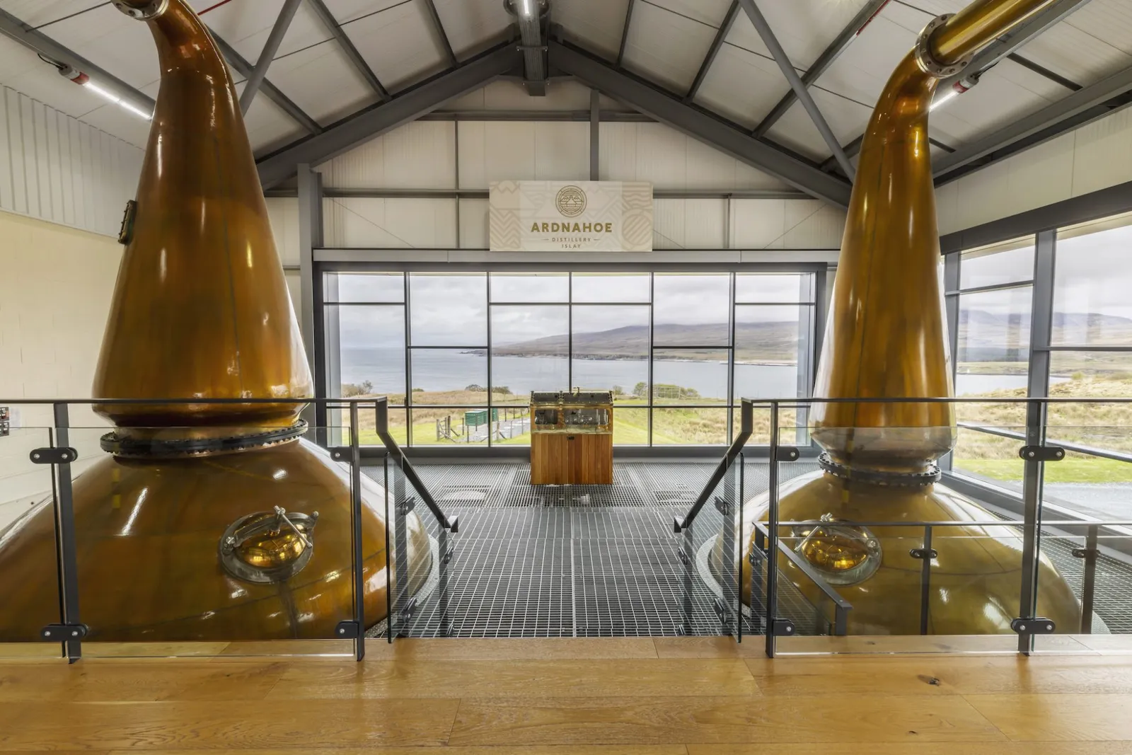Interior view of Ardnahoe Distillery showing two large copper pot stills framing a large glass window overlooking the coastal scenery of Islay