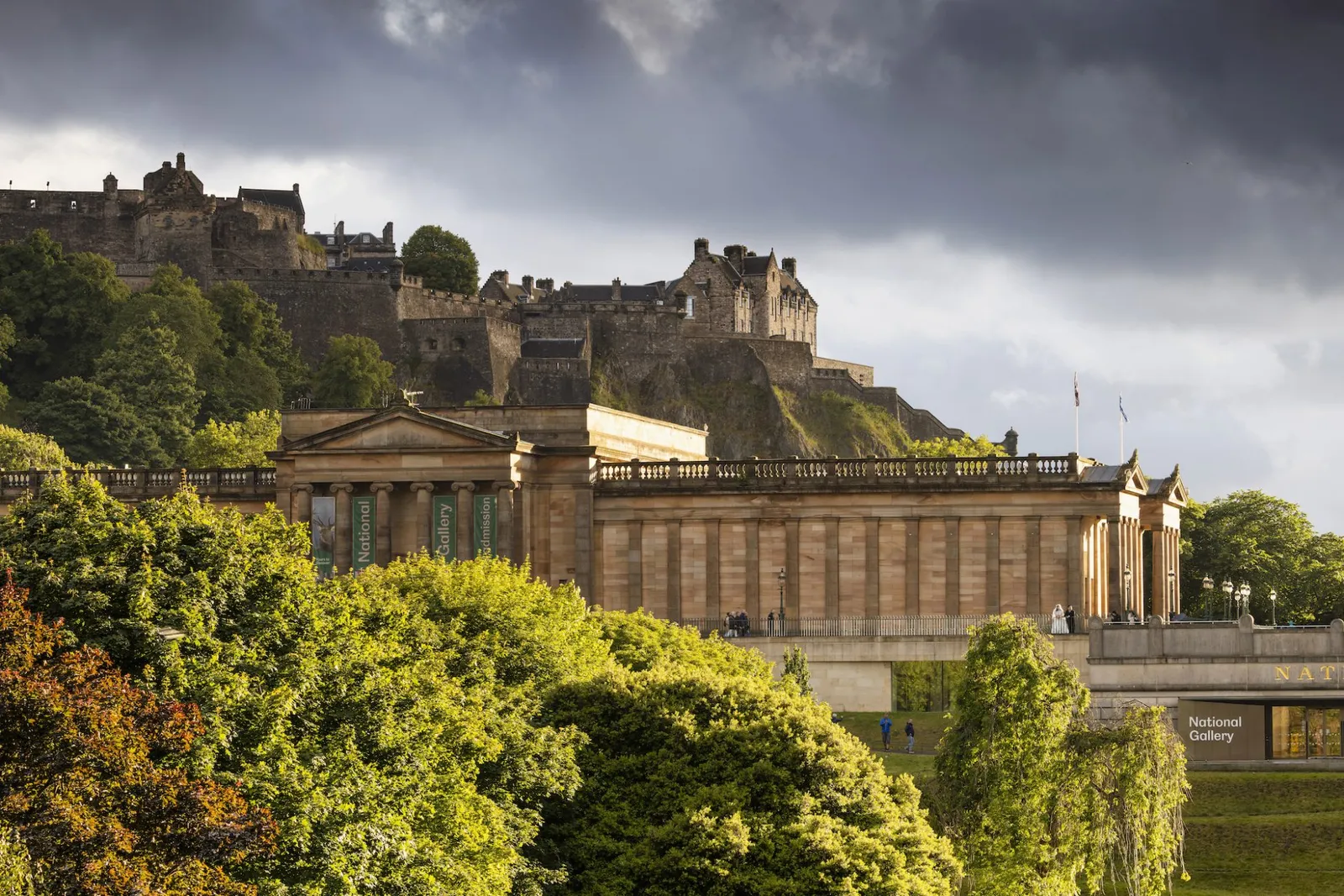 The National Gallery of Scotland in the foreground with classical columns, backed by Edinburgh Castle atop Castle Rock under dramatic clouds