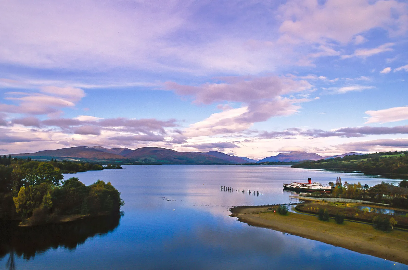 Scenic wide view of Loch Lomond with a steamship docked near the shore and hills under a soft blue and purple sky