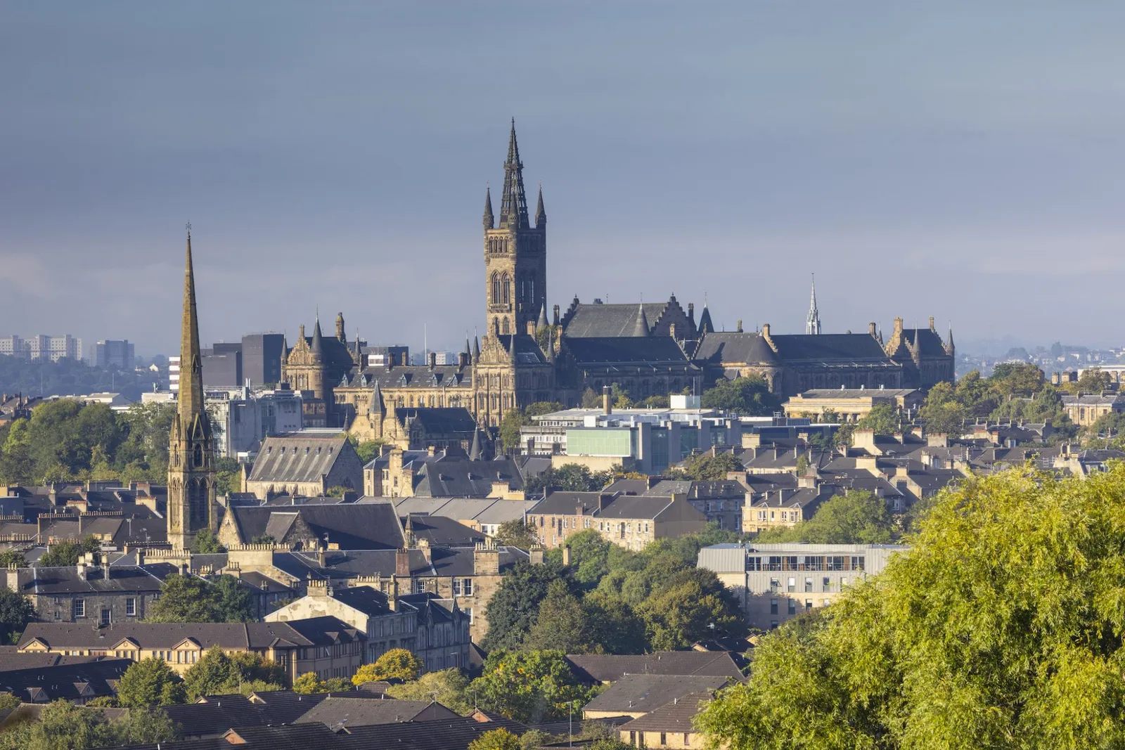 Elevated cityscape view of the Glasgow skyline featuring the Gothic spire of the University of Glasgow main building surrounded by historic architecture and leafy trees under a clear sky